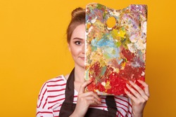 Close up portrait of young woman painter against yellow studio wall, draws picture, lady wearing casual striped shirt and brown apron, girl artist covers half of her face with palette of colours.