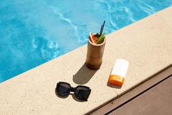 Flat lay of cocktail in wooden container, black fashionable sunglasses and sun protection cream in white bottle, making up composition for rest during vacation. Things are situated near swimming pool.