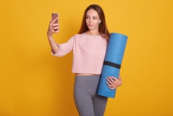 Image of young and active dark haired woman posing with blue yoga mat and makes selfie via phone in studio isolated over yellow background, lady wearig stylish sportwear. Fitness and diet concept.