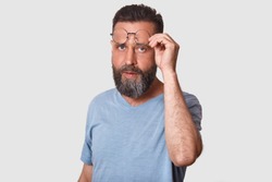 Closeup portrait of handsome young bearded man with glasses, wearing casual clothing, standing over white studio background, raising his eyewear questioningly, doubts and looking directly at camera.