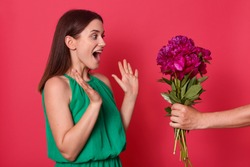 Studio shot of impressed sweet good looking brunette raising her hands, opening eyes and mouth widely, being surprised by present of bright peonies, unknown hand holding flowers. Present concept.