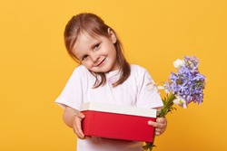 Horizontal shot of shy sweet lovely little girl smiling sincerely, looking directly at camera, holding red and white box, blue flowers, giving present, standing isolated over yellow background.