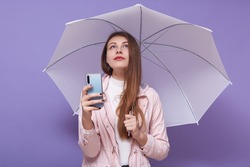 Portrait of pensive good looking tender female looking up, holding smartphone and pink umbrella, using her device, taking photos, having long hair, staying alone, being dreamy. Youth concept.