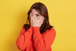 Picture of shy brunette teenage girl covering face with hands and peeping through her fingers at camera. Young attractive female hiding her face being frightened, scared or ashamed, wears casually.
