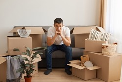 Sad upset man sitting on sofa surrounded with pile of packages, looking at camera with pout lips, doesn't want to move from his favorite apartment or doesn't want to unpack boxes with belongings.