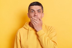 Shocked man wearing casual style hoodie, posing isolated over yellow background, covering mouth with hand to keep silent, afraid to say secret, looking with intimidated expression