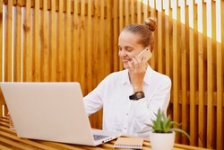 Image of attractive adorable young adult woman talking on mobile phone working on laptop and having communication with her business partner, wearing white shirt.