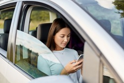 Side view of attractive adorable woman with dark hair wearing white shirt sitting in her car using her cell phone, looking at display of smart phone.