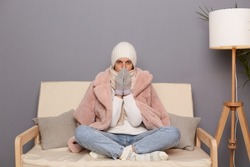 Portrait of frozen beautiful woman wearing warm clothes and wrapped in scarf sitting on sofa in room, trying to get warm in cold apartment, looking at camera.