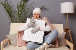 Portrait of Caucasian woman in coat and hat sit in cold living room embracing electric heating to get warm and using cell phone, call to service, broken central heating.