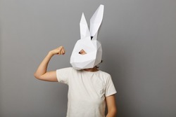 Indoor shot of powerful strong woman wearing white t shirt and paper bunny mask, raised her arm and showing her biceps and her power, standing isolated over gray background.