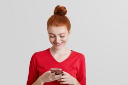 Attractive ginger young female in red clothes holds smart phone, types text messages, surfes social networks or online webpage, connected to wireless internet, isolated over white background
