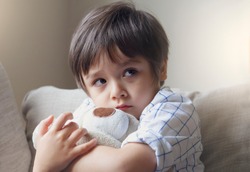 Dramatic portrait of little boy sitting on sofa and cuddling teddy bear with scared face,Unhappy Child sitting alone and looking out with worrying face,Toddler boy on corner punishment sitting.