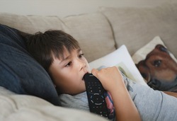 Happy mixed race kid lying on sofa biting remote contole while watching TV, Child boy laying on couch relaxing at home after back from school.