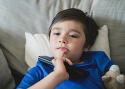Happy mixed race boy putting finger on his chin while watching TV, Candid shot Youg kid lying on sofa holding remote contro, Positive child relaxing at home on weekend