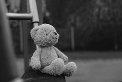 Lost teddy bear sitting on swing at playground in gloomy day, Lonely and sad face brown bear doll sitting alone in the park, lost toy or Loneliness concept, International missing Children day