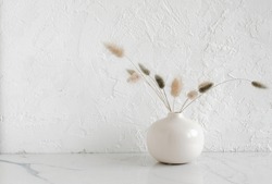 Modern beige ceramic vase with dry fluffy flowers on marble table near a white textured wall. Copy space.Minimal Scandinavian interior. Neutral trendy colors interior decoration .
