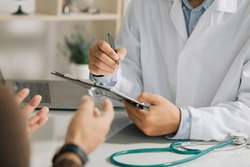 A professional physician in a white medical uniform talks to discuss results or symptoms and gives a recommendation to a male patient and signs a medical paper at an appointment visit in the clinic.
