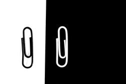 A white-clipped paperclip sits on a black surface and another black paperclip sits on a white surface - black and white concept with paperclips - stark contrasts and differences as background
