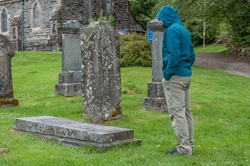 Man looking at a grave in ancient Scottish Balquhidder Parish church cemetery on a rainy day. Concept: visit to mysterious and evocative places in Scotland