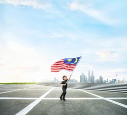 Happy boy running while holding Malaysia flag on open space with Kuala Lumpur cityscape.