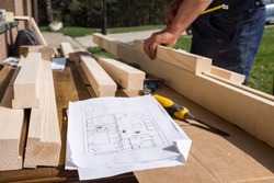 Carpenter working with technical blueprint drawing construction paper lying on outdoor workshop desk,surrounded with carpentry tools & wood,furniture making pastime,cabin or house renovation process