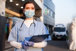 Young female EMS key worker doctor in front of healthcare ICU facility,wearing protective PPE face mask equipment,holding medical lab patient health check form,UK US COVID-19 pandemic outbreak crisis 