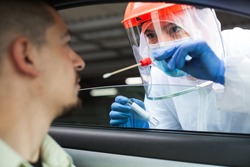 Medical NHS UK worker performing drive-thru COVID-19 check,taking nasal swab specimen sample from male patient through car window,PCR diagnostic for Coronavirus presence,doctor in PPE holding test kit