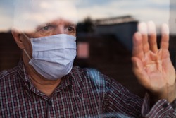 Portrait of elderly senior citizen wearing face mask looking through room window,Coronavirus COVID-19 pandemic outbreak nursing home crisis,high mortality rate and death cases among older population