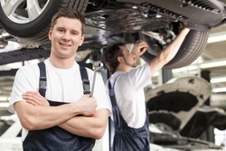 Mechanics at work shop. Confident young mechanic standing with his arms crossed and smiling at camera while another one working on the background