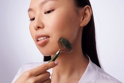 Young asian girl doing massage with stone roller. Woman face massage. Woman doing Guasha face drainage massage with jade stone roller. Cosmetology and self-care. Ideal skin.