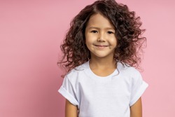Closeup shot of charming caucasian little girl in white t shirt, with friendly, kind look looking at camera with cute smile, posing against pink wall. Happy childhood, childish innocence, kids concept