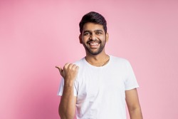 Waist-up shot of cheerful carefree indian man with stubble, wearing white t shirt, pointing left with thumb, laughing, looking at camera, standing over pink background. Advertising concept.