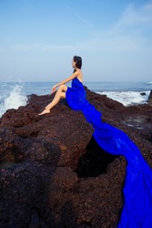 fashion pretty female model posing on a beach with rocks in a long butterfly chameleon dress waterfall of skirt plume train.sensual perfume with a beautiful and young brunette woman wave tropical sand
