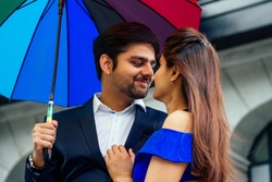 indian woman in long evening blue dress spinding romantic time together with lover handsome boyfriend under multi-colored rainbow umbrella europe urban downtown city.