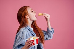 cute red long foxy haired ginger woman holding packaging cardboard box with french fries in pink studio