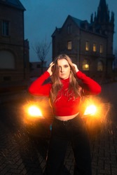 Young blonde woman with a red jersey and black trousers sit-in a red car next to a old castle at night.