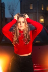 Young blonde woman with a red jersey and black trousers sit-in a red car next to a old castle at night.