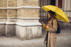 Young beautiful woman using a smartphone and holding a yellow umbrella while walking in the city on a rainy day
