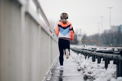 Rear backside view of a woman jogging on a bridge with snow on cold winter day. Urban city jogger. Woman in sportswear running on a bridge while snowing.