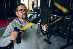 Fat middle aged man training in the gym while using trx suspension. Middle aged chubby man trying to reduce weight. Overweight.