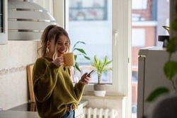 Shot of a young woman drinking tea and using smartphone in her kitchen while getting ready to go to work. Watching videos or messaging with friends while enjoying morning at home.