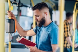 Young cheerful man reading a popular fiction book during his ride and holding onto the bar while standing in a bus. Handsome bearded man taking bus to work. Urban public transportation concept.