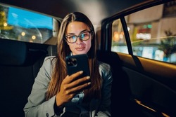 Beautiful business woman using smartphone in a car at night. Busy female working on a mobile phone in the back seat of a taxi. Bokeh and neon city lights in the background.