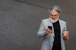 Portrait of a senior business executive man smiling and using mobile phone and drinking coffee. A elderly businessman standing on the street surrounded by buildings and using smartphone. Copy space.