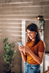 Beautiful woman checking social media while using smartphone and airpods at home. Smiling young female using mobile phone app and playing game, shopping online or reading news. Copy space.