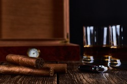 Still life with three cuban cigars, two glasses of whiskey or rum, lighter and wooden box with hygrometer. Old wooden table top and black background. Space for your text.