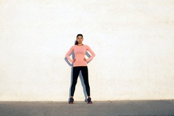 Fit young woman standing holding her arms on her hips, on the white concrete wall background, looking forward, wearing black sport tights and pink long sleeve shirt
