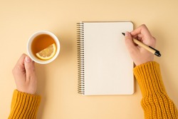 First person top view photo of female hands in yellow pullover writing in spiral planner and holding white cup of tea with lemon slice on isolated pastel orange background with copyspace