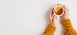 First person top view photo of female hands in yellow sweater touching white cup of tea with lemon slice on isolated white background with copyspace